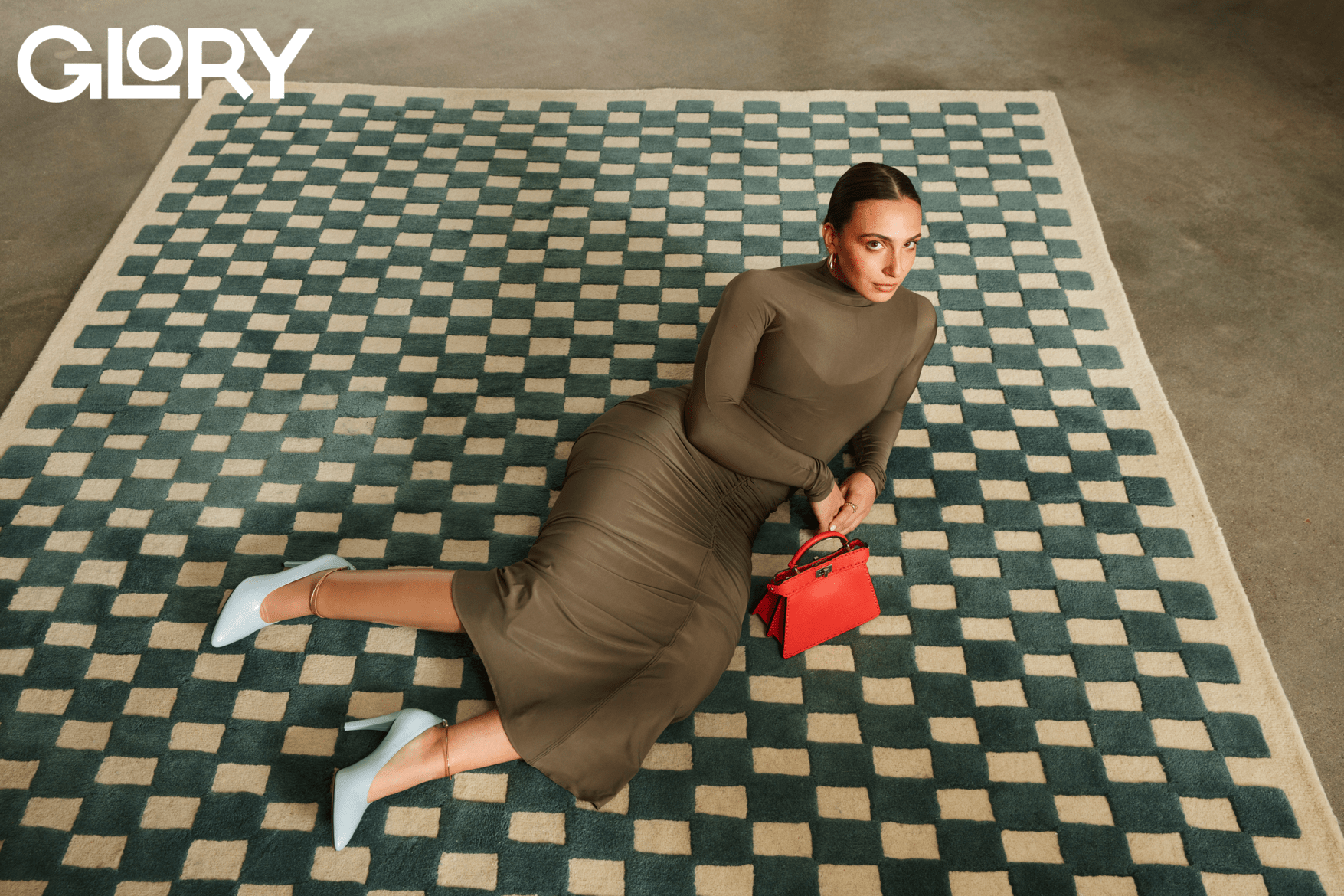 Marissa Papaconstantinou wearing a sheer taupe dress lying on a checkered rug with a red leather purse.