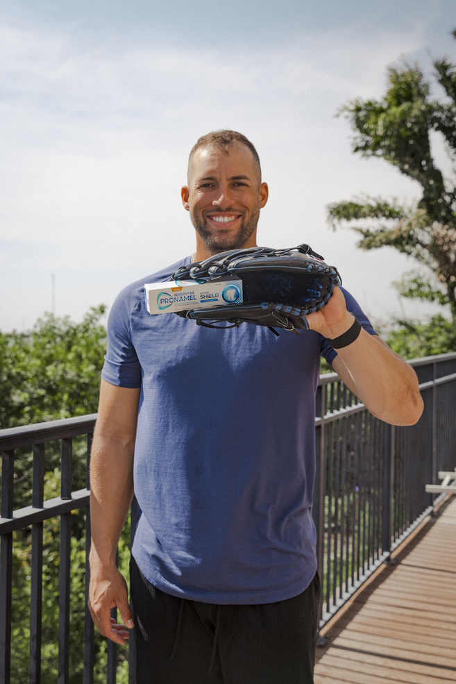 George Springer wearing a blue t-shirt on a sunny balcony and holding a pronamel package with a black baseball mitt and smiling into the camera.