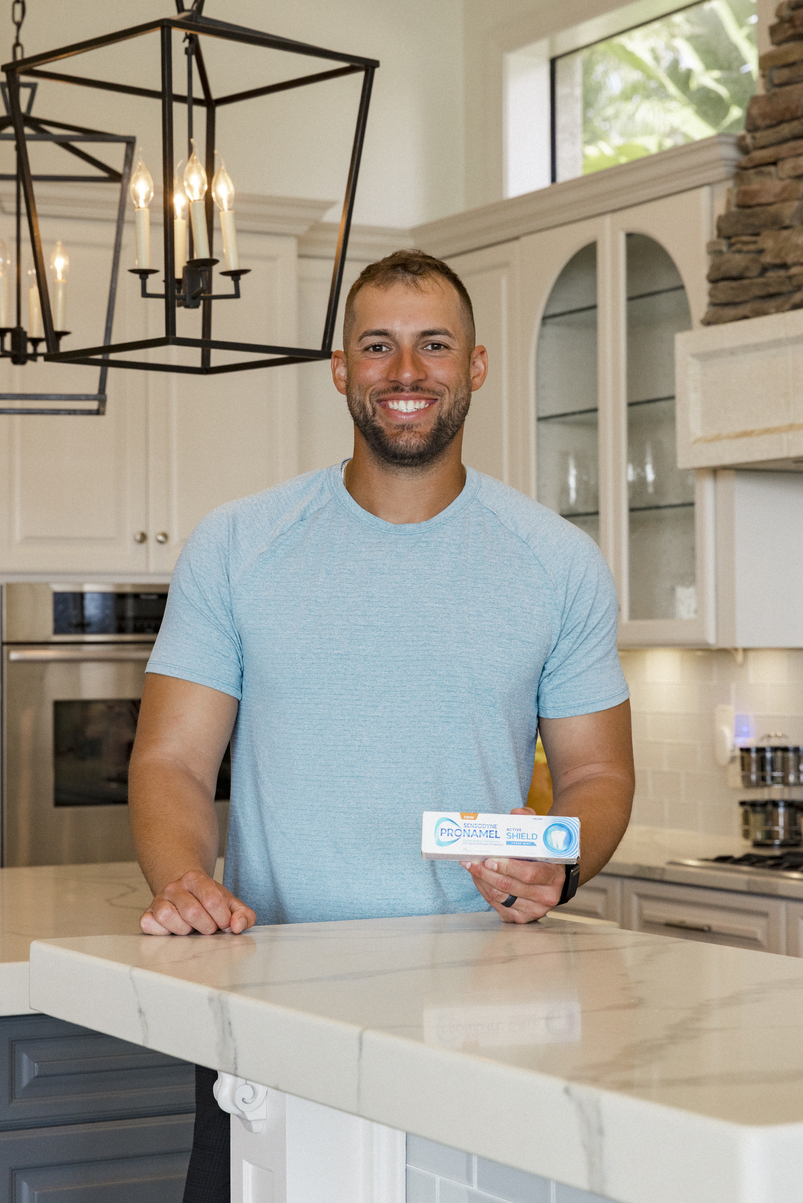 George Springer wearing a blue t-shirt in a kitchen and holding a pronamel package and smiling into the camera.